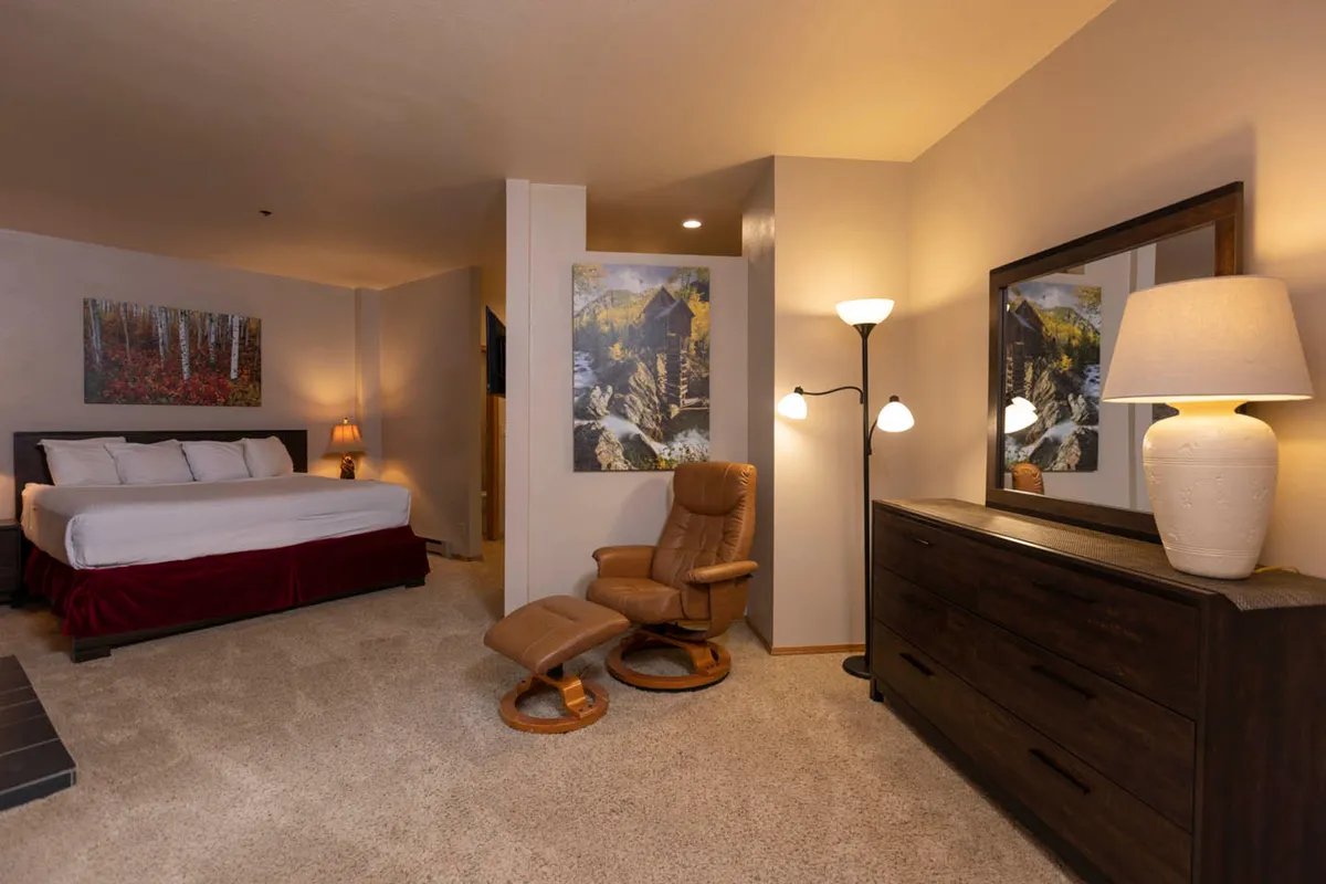 A serene bedroom featuring a plush bed, an armchair, a dresser, soft lighting from a bedside lamp, and tranquil artwork. A large window frames scenic mountain views, filling the room with natural light.
