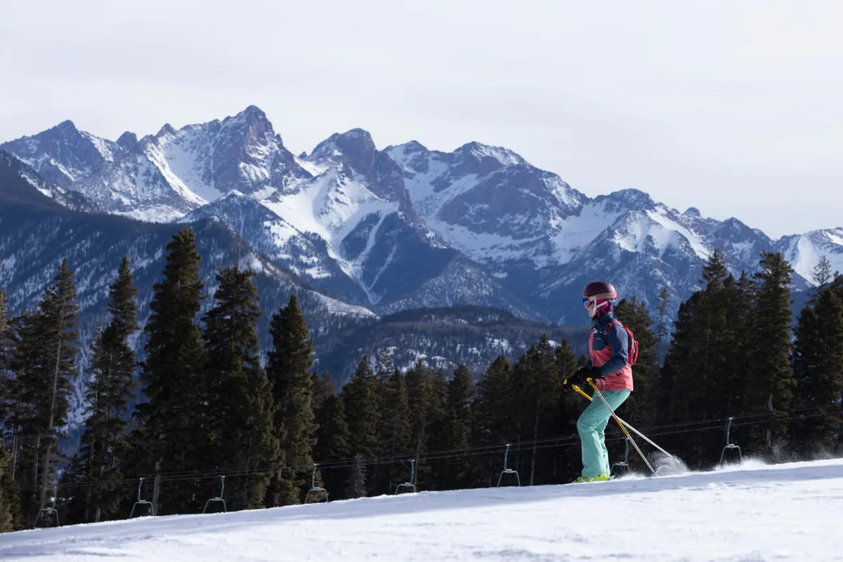 A skier carves down a snow-covered slope with a striking mountain range as the backdrop. The scene is dynamic and full of energy, capturing the thrill of downhill skiing and the natural alpine beauty that defines Purgatory Resort in winter.