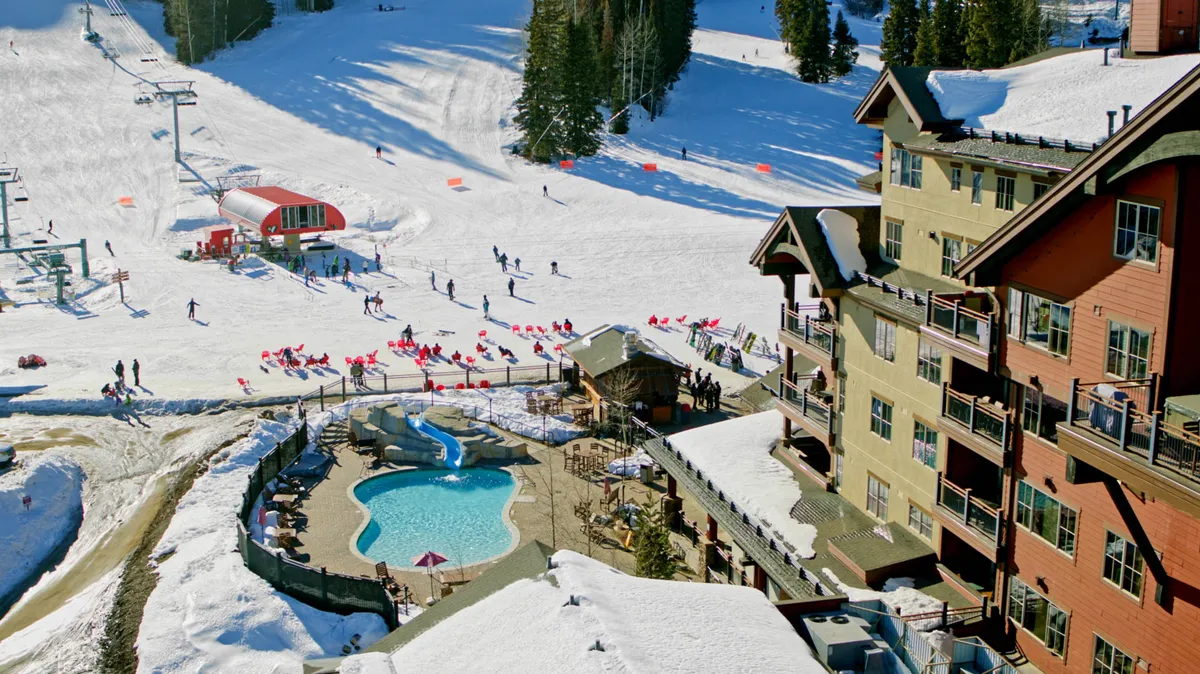This image captures the vibrant heated outdoor pool area with the backdrop of ski slopes, a ski lift, and surrounding trees. The pool is directly adjacent to lodge architecture, illustrating how recreation and relaxation blend seamlessly at the resort.