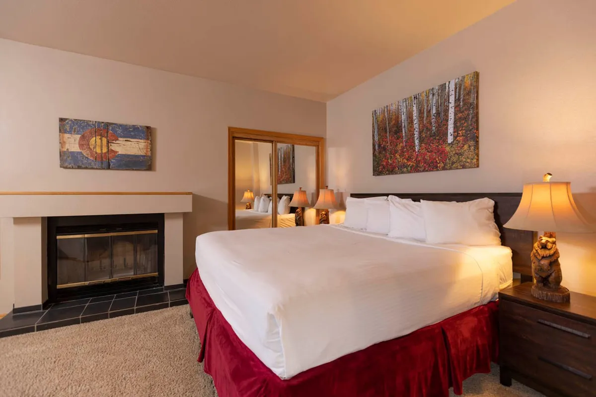 A welcoming bedroom with a bed dressed in crisp white linens, a warm fireplace, and tasteful decorative paintings. The inviting ambiance is perfect for unwinding after a day outdoors.