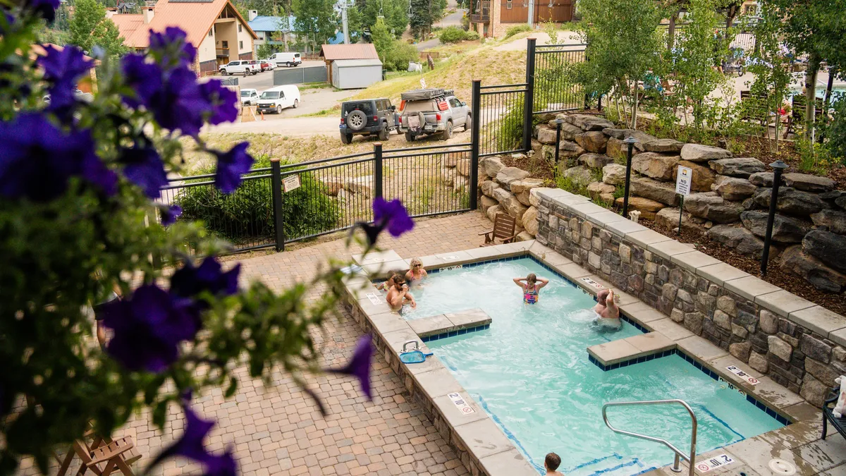 A cheerful outdoor hot tub scene surrounded by lush greenery and stone walls, with guests socializing and relaxing amid the mountain air. The inviting space evokes relaxation after a day of outdoor activities.