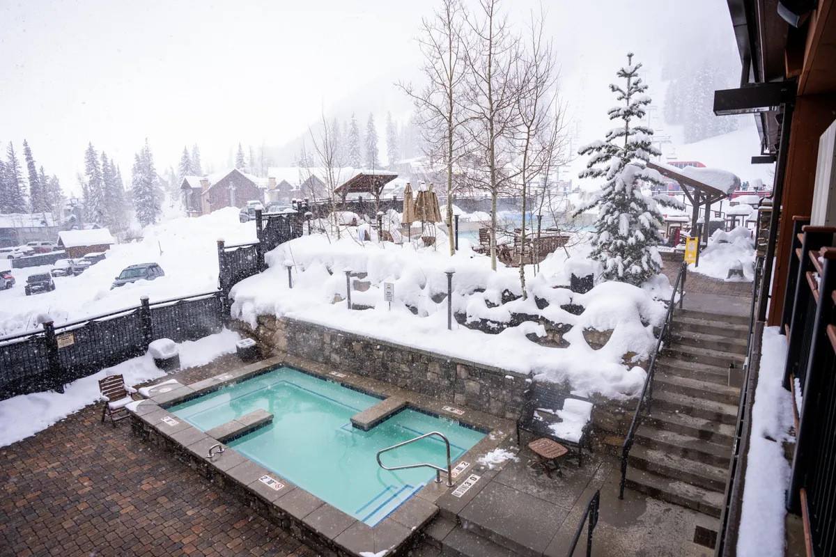 This image showcases an inviting outdoor hot tub set amid a snow-covered landscape, surrounded by trees and with stairs leading to the tub. The tranquil, wintry setting is ideal for relaxation and socializing, evoking a peaceful resort experience.