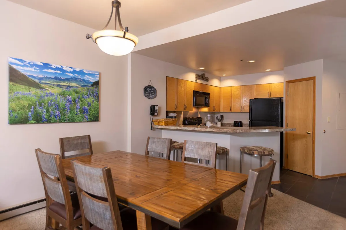 This image showcases a welcoming interior with a dining table and kitchen area visible. The open layout allows for easy interaction between those cooking and those seated at the table. Landscape artwork and natural light contribute to a warm, inviting atmosphere, hinting at scenic views beyond.