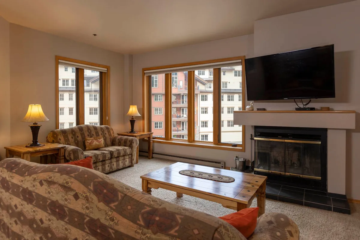 A cozy living room featuring large windows with scenic mountain views, a crackling fireplace as a focal point, a comfortable sofa, and a wooden coffee table arranged for conversation and relaxation. The soft lighting and inviting seating create a space ideal for gathering after a day outdoors.