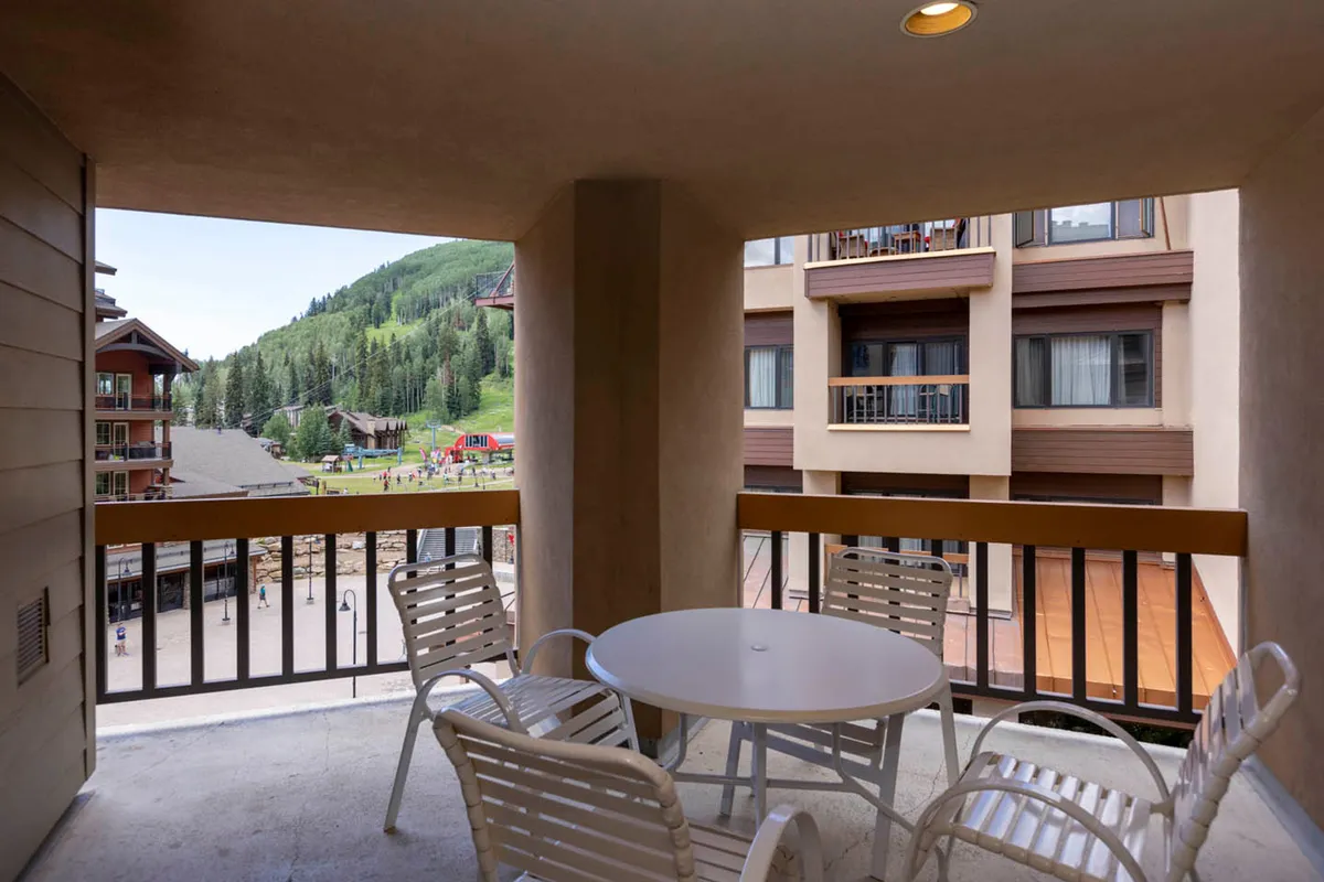 A serene balcony scene overlooks the mountains and resort architecture, with comfortable seating suggesting moments of relaxation or socializing with family. The image evokes the pleasure of morning coffee or evening drinks enjoyed while watching children play below.
