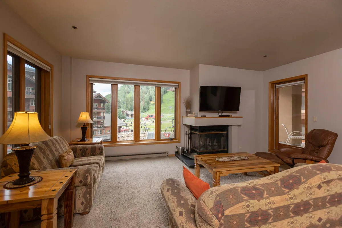 The image showcases a warm, inviting living room with plush seating, a crackling wood-burning fireplace, and expansive windows revealing stunning mountain views. The scene exudes comfort and relaxation, with natural light pouring in and a perfect spot for gathering after a day of adventure.
